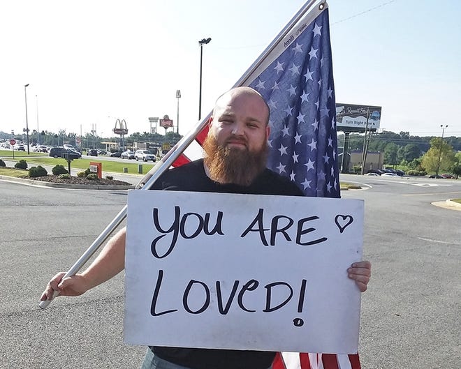 In this Aug. 12, 2019, photo Caleb Snell, 27, holds a sign in Athens, Ala. Snell uses the signs at area intersections in Athens, Huntsville and Decatur to remind others they are loved. (Lora Scripps/Athens News Courier via AP)