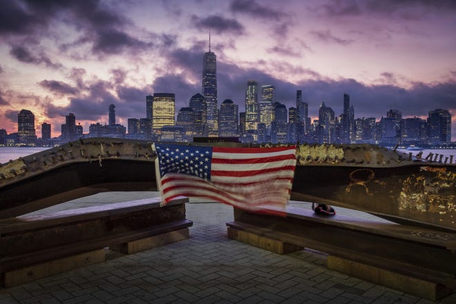 A U.S. flag hanging from a steel girder, damaged in the Sept. 11, 2001 attacks on the World Trade Center, blows in the breeze at a memorial in Jersey City, N.J., as the sun rises behind One World Trade Center building and the re-developed area where the Twin Towers of World Trade Center once stood in New York City on the 18th anniversary of the attacks. [AP Photo/J. David Ake]