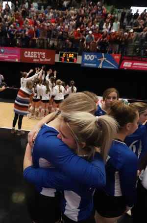 Holy Trinity's Avery Hopper, left, and Taylor Crabtree hug after they lost their semifinal match of the Class 1A Iowa state volleyball tournament to Wapsie Valley, who celebrate with their fans in the background, Thursday in Cedar Rapids. [John Gaines/thehawkeye.com]