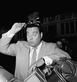 Television comedian Jackie Gleason dons a party hat as he awaits a customs inspection following his arrival from Europe, Aug. 4, 1954, New York. [John Lent/The Associated Press]