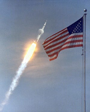 The American flag heralds the flight of Apollo 11, the first Lunar landing mission. The Apollo 11 Saturn V space vehicle lifted off with astronauts Neil A. Armstrong, Michael Collins and Edwin E. Aldrin, Jr., at 9:32 a.m. EDT July 16, 1969, from Kennedy Space Center's Launch Complex 39A. During the planned eight-day mission, Armstrong and Aldrin will descend in a lunar module to the Moon's surface while Collins orbits overhead in the Command Module. The two astronauts are to spend 22 hours on the Moon, including two and one-half hours outside the lunar module. They will gather samples of lunar material and will deploy scientific experiments which will transmit data about the lunar environment. They will rejoin Collins in the Command Module for the return trip to Earth.