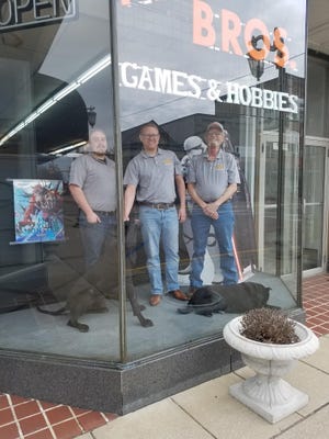 Customers have kept M.A.D. Bros. Games & Hobbies afloat by paying for games before they are available, according to co-owner Todd Smith (center), shown in the front window of the downtown New Philadelphia store with employee Robert Kloha (left), father Mark Smith (right), dogs Maisy (left) and Skyy. (TimesReporter.com / Nancy Molnar)