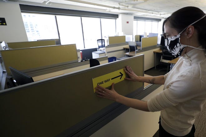 HOLD FOR STORY BY KELVIN CHAN — In this Thursday, May 7, 2020 photo, Interior Designer Stephanie Jones at the design firm Bergmeyer, applies a safe distancing reminder indicating one way foot traffic to a cubicle at the firms offices, in Boston. Out of concern for the coronavirus Bergmeyer is restructuring the way its workspace is used, including reinstalling dividers on 85 desks at its Boston office that had been removed over the years. (AP Photo/Steven Senne)