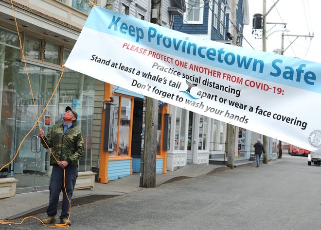 Brian Cowing, a foreman with the Provincetown Department of Public Works, hangs a banner across Commercial Street near Town Hall on Friday offering safety reminders about the coronavirus. A public health working group has recommended curbing many popular activities this summer. [Merrily Cassidy/Cape Cod Times]