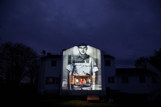 An image of veteran Charles Lowell is projected onto the home he shared with his wife, Alice, for 30 years as she stands at left with her daughter, Susan Kenney, in Hardwick, Mass. Saturday, May 2, 2020. Lowell, a U.S. Air Force veteran and resident of the Soldier's Home in Holyoke, Mass., died from the COVID-19 virus at the age of 78. Seeking to capture moments of private mourning at a time of global isolation, the photographer used a projector to cast large images of veterans on to the homes as their loved ones are struggling to honor them during a lockdown that has sidelined many funeral traditions. (AP Photo/David Goldman)
