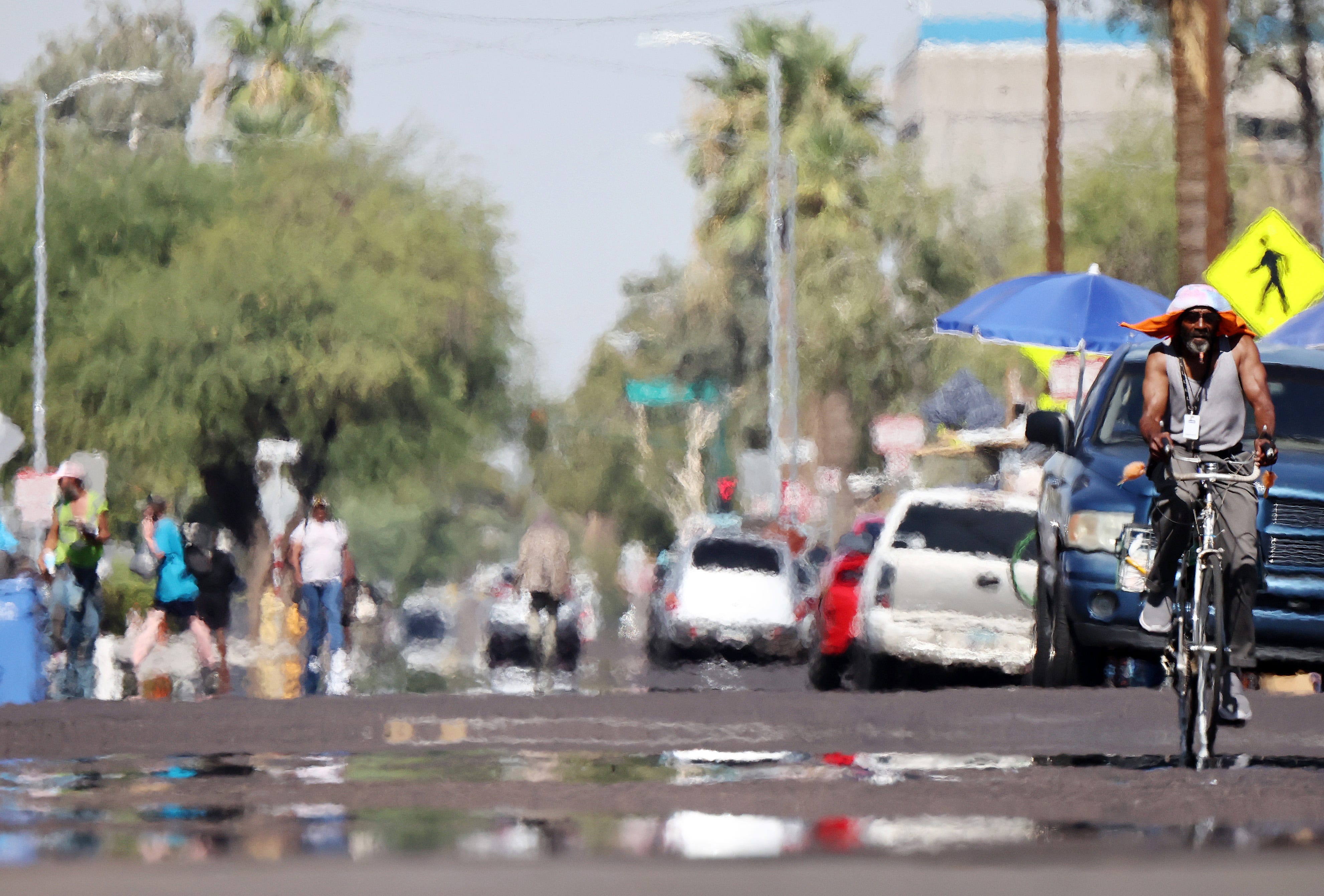 A person rides a bicycle as heat waves shimmer, causing visual distortion, as people walk in the 'The Zone', Phoenix's largest homeless encampment, amid the city's worst heat wave on record on July 25, 2023 in Phoenix, Arizona. While Phoenix endures periods of extreme heat every year, today is predicted to mark the 26th straight day of temperatures reaching 110 degrees or higher.