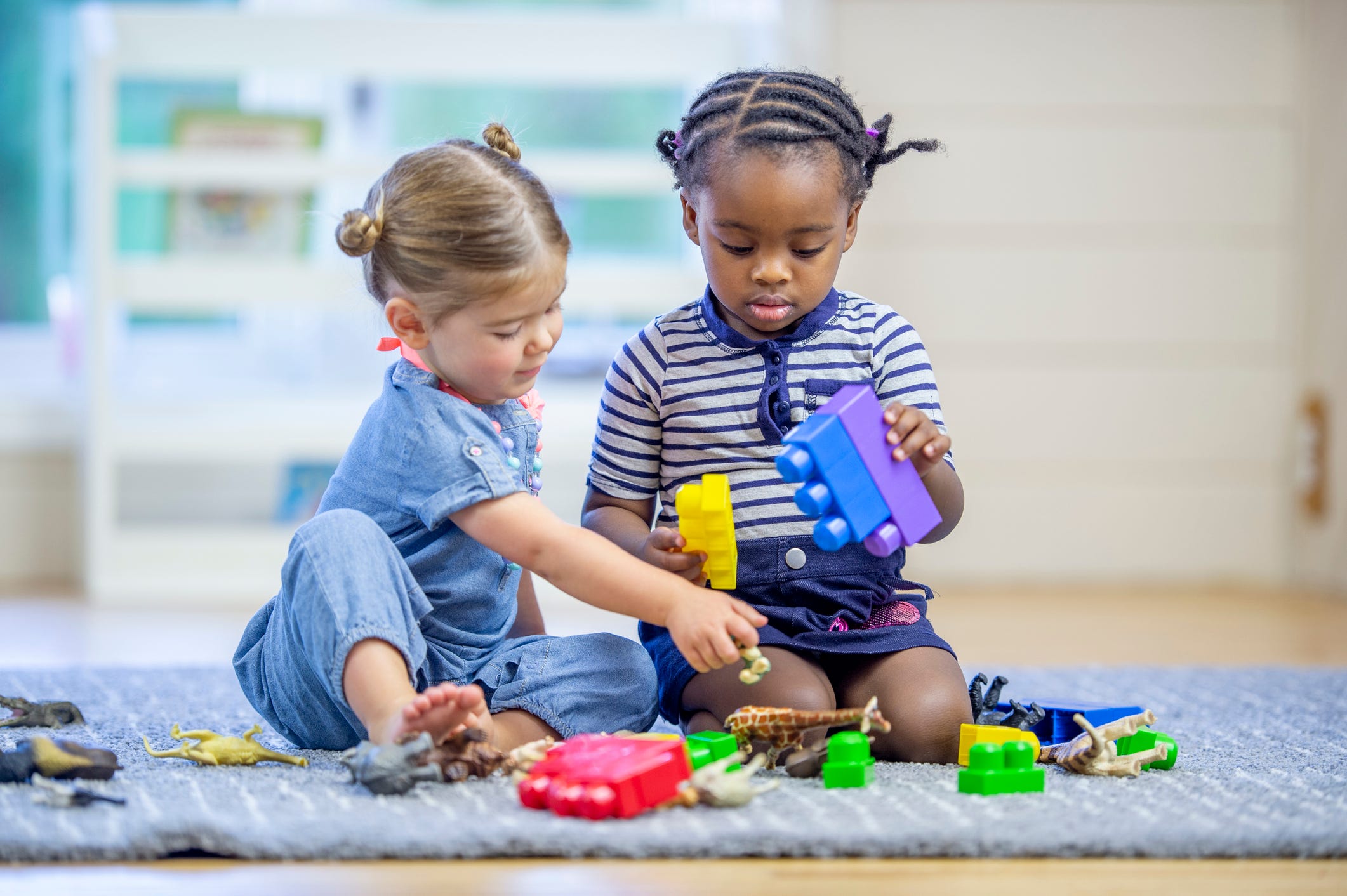 Two young toddlers sit on the carpet in a home daycare as they play together with toy animals and blocks.