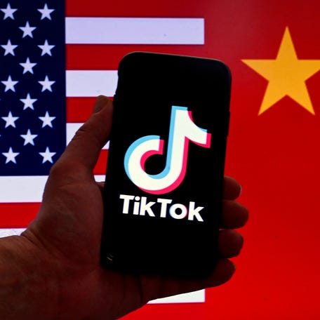 The social media application logo for TikTok is displayed on the screen of an iPhone in front of a US flag and Chinese flag background in Washington, DC.