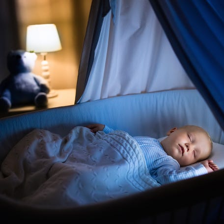 Baby pictured sleeping in blue bassinet with canopy at night.
