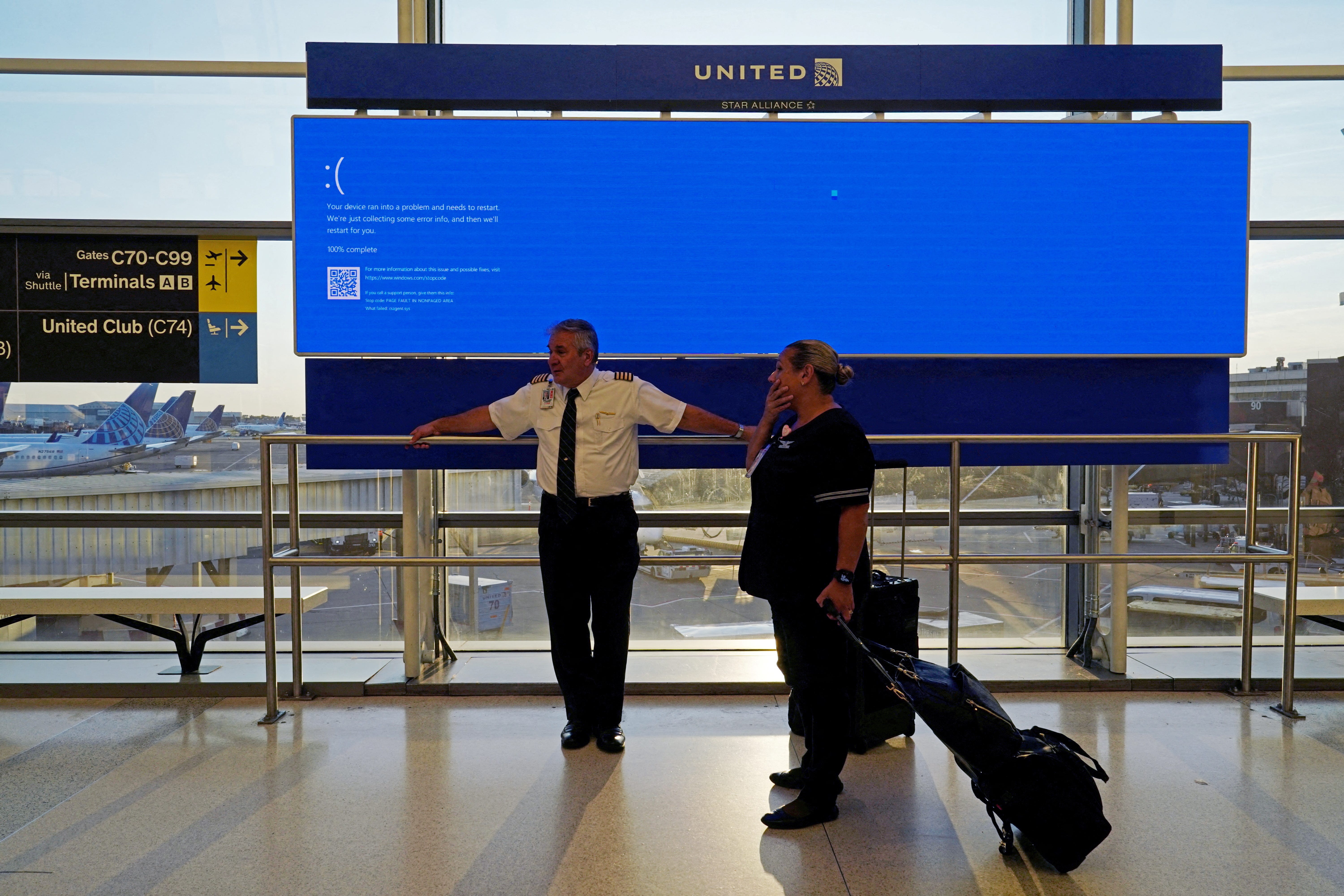 United Airlines employees wait by a departures monitor displaying a blue error screen, also known as the 