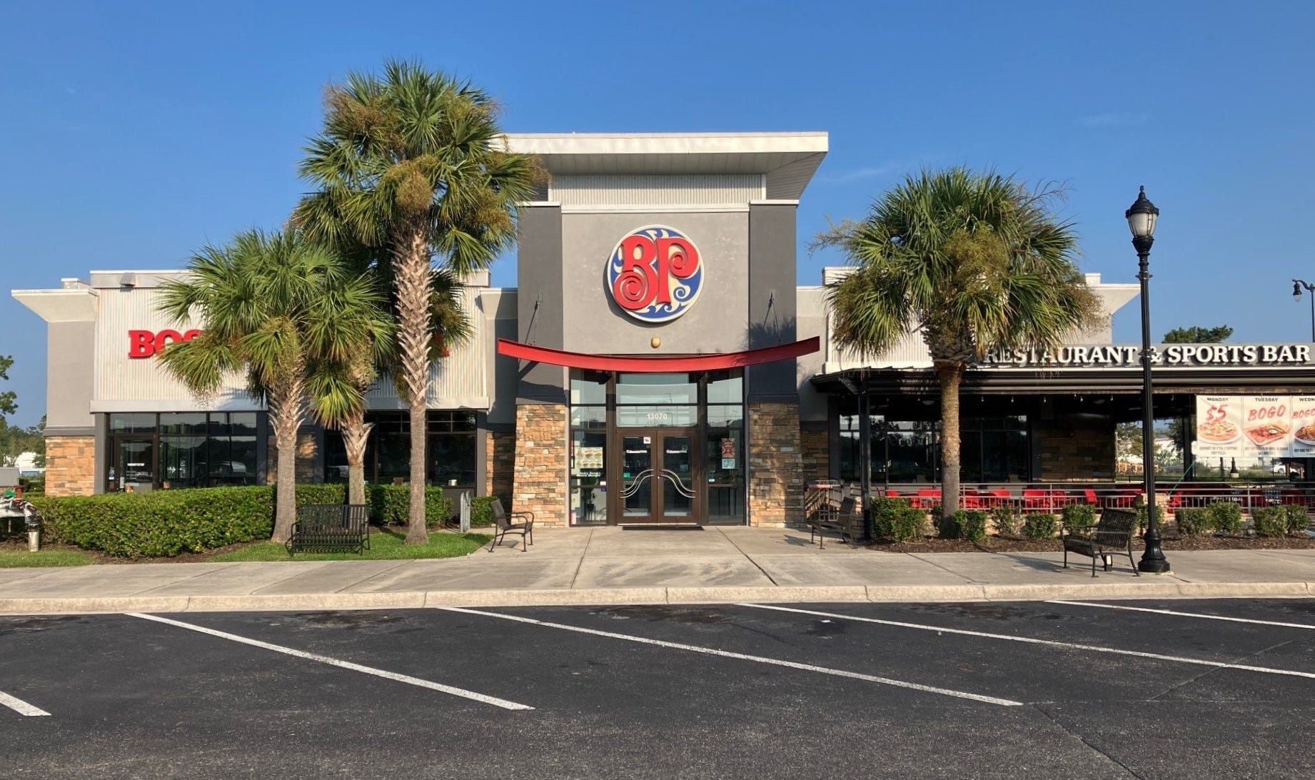 The image shows the exterior of a restaurant and sports bar with a modern design, featuring a central entrance framed by stone accents and a gray façade. Above the entrance is a circular logo with the letters 'BP' in red against a blue and white background, and a red curved awning extends over the door. On either side of the entrance are palm trees, benches, and neatly trimmed hedges, while a black lamp post stands to the right near an outdoor seating area with red chairs, partially visible under a covered patio. The parking lot in front is empty, and the sky is clear with no clouds.