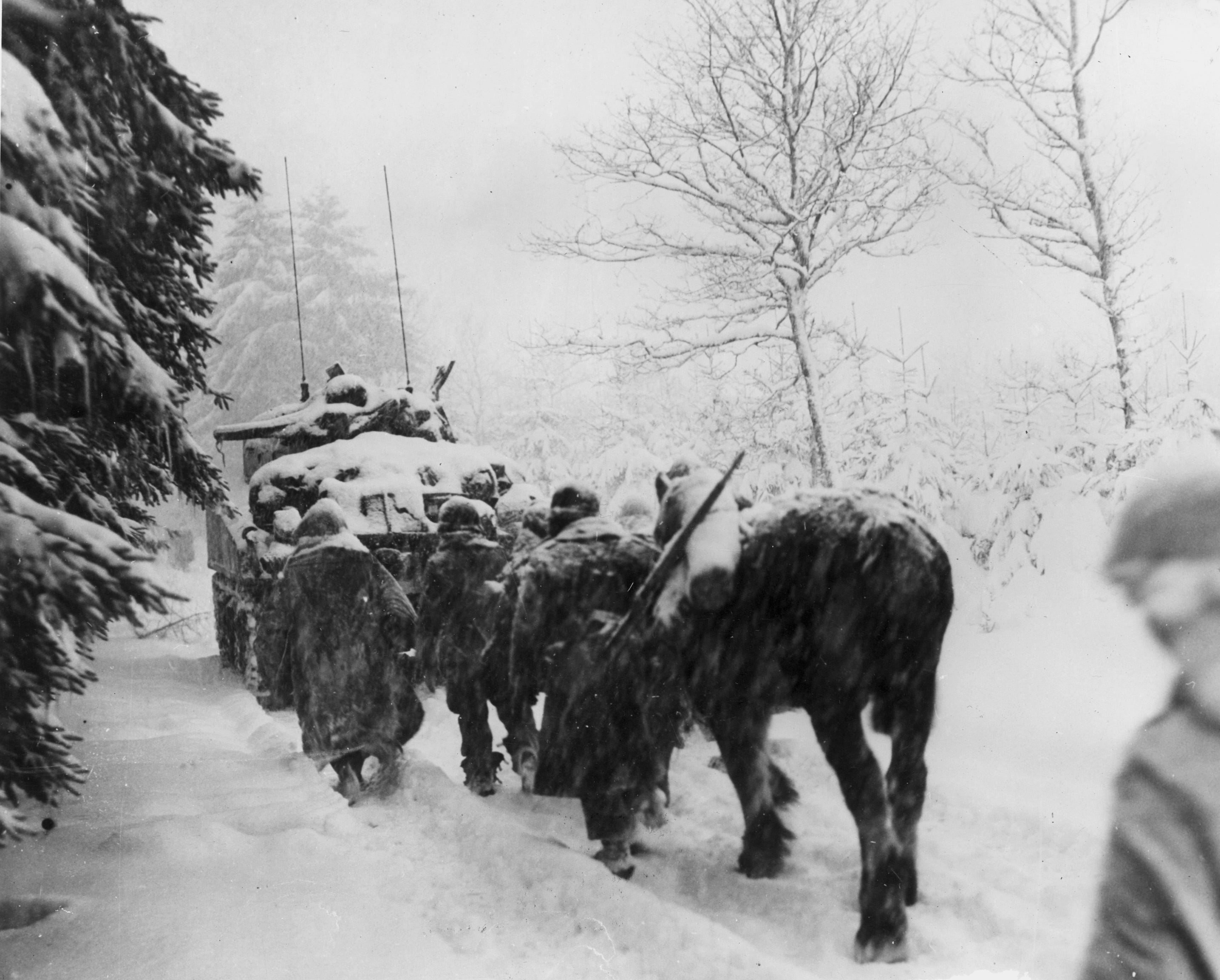 Members of the American 82nd Airborne Division trudging through the snow behind a tank during the Battle of the Bulge.