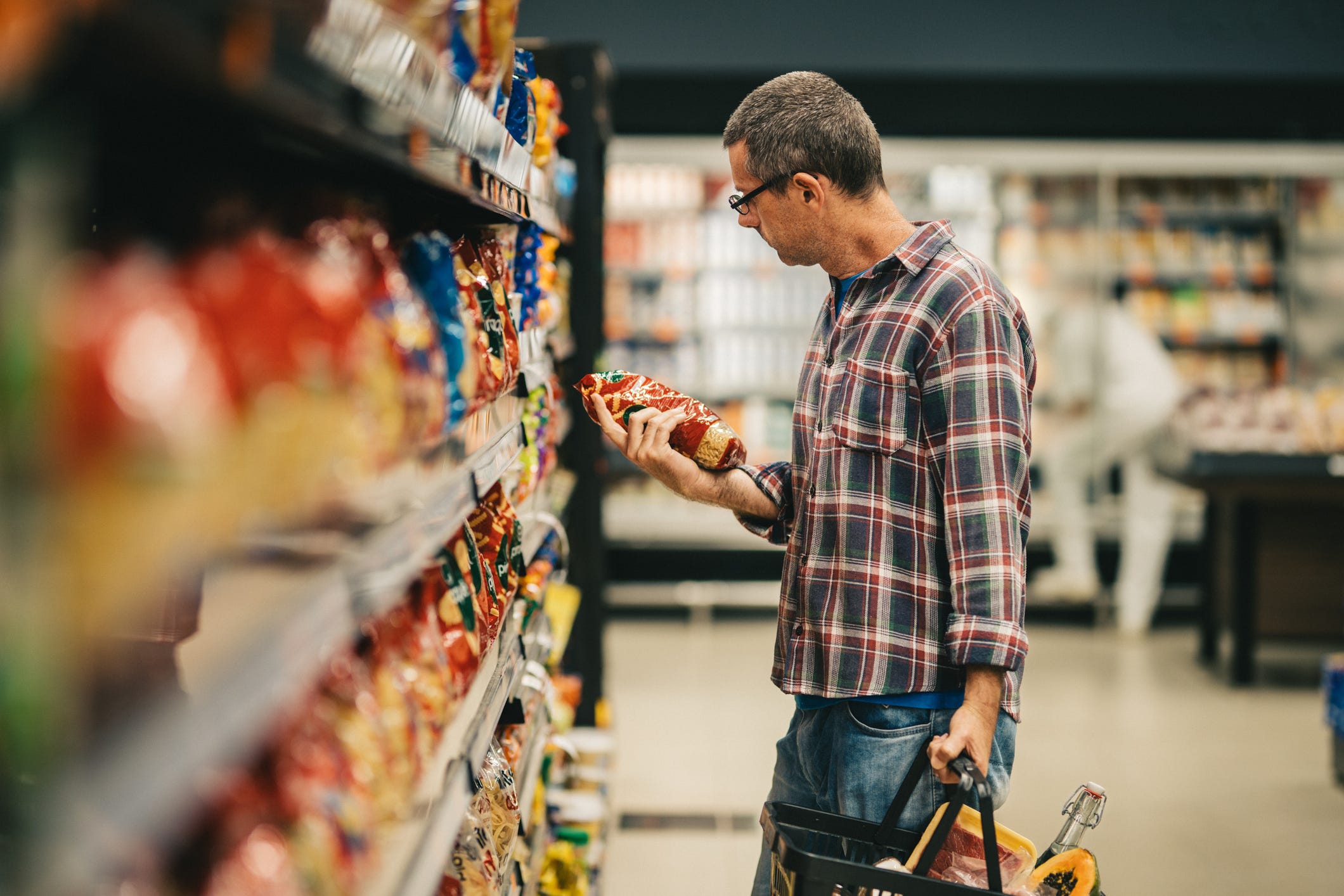 A man stands in a supermarket aisle and reads the label on a bag of pasta while grocery shopping.