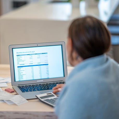 A young woman sits at her kitchen table with her laptop open in front of her with a spreadsheet on it, bills on the counter and a calculator at her fingertips as she tries to budget.