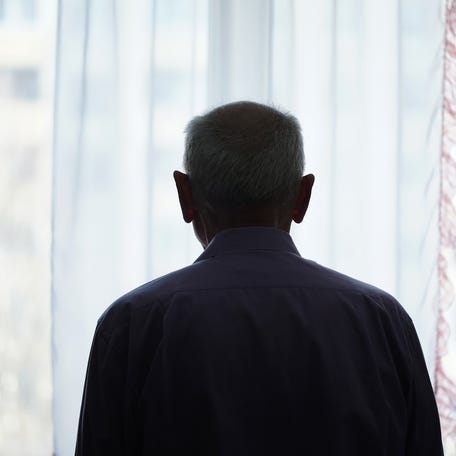 Silhouette of retired man looking through window with transparent curtain standing at home rear view. Loneliness and old human care concept.