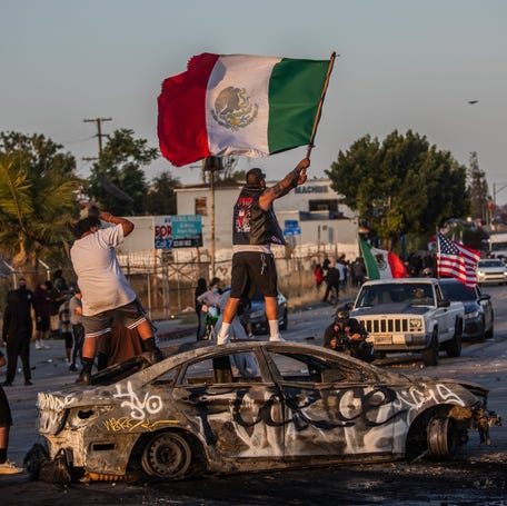 A protester stands on a burned car holding a Mexican flag on June 7, 2025, in Paramount, California.