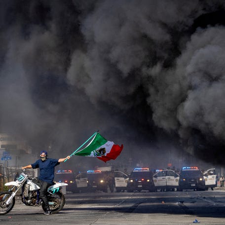 A man on a motorcycle waves a Mexican flag as smoke rises from a burning car on Atlantic Boulevard, during a standoff by protesters and law enforcement, following multiple detentions by Immigration and Customs Enforcement (ICE), in the Los Angeles County city of Compton, California, U.S., June 7, 2025.