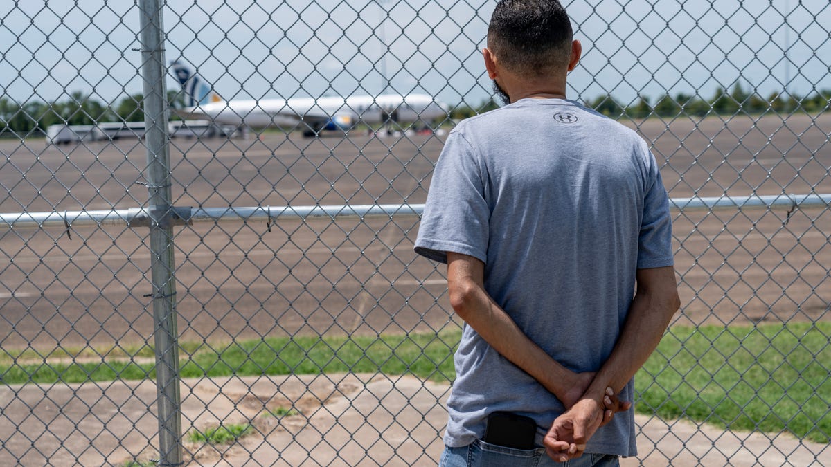 Sam Ziden watches behind chain link, hoping to catch a glimpse of his brother in a group of migrants being loaded onto an airplane at the Alexandria Staging Facility in Alexandria, Louisiana on June 11, 2025.