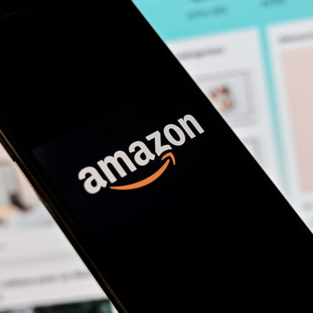 A person poses with a smartphone showing an Amazon logo in front of a computer screen displaying the home page of the Amazon sales website.