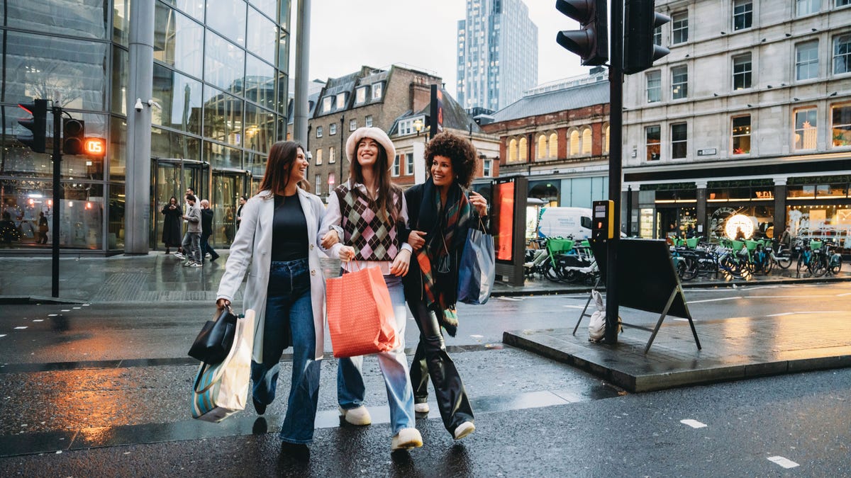 Young women walk arm in arm on a crosswalk in London carrying shopping bags.