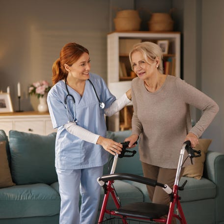 Smiling nurse supports senior woman learning to walk with a walker in her living room.