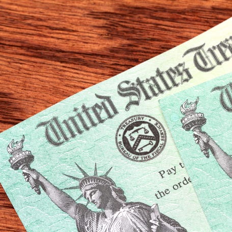 Two US Treasury checks of the type used to pay tax refunds as well as Medicare, Social Security and welfare benefits are partially visible on a wood desk background. Checks show the Treasury seal and image of the Statue of Liberty.