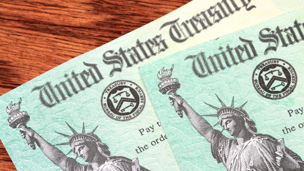 Two US Treasury checks of the type used to pay tax refunds as well as Medicare, Social Security and welfare benefits are partially visible on a wood desk background. Checks show the Treasury seal and image of the Statue of Liberty.