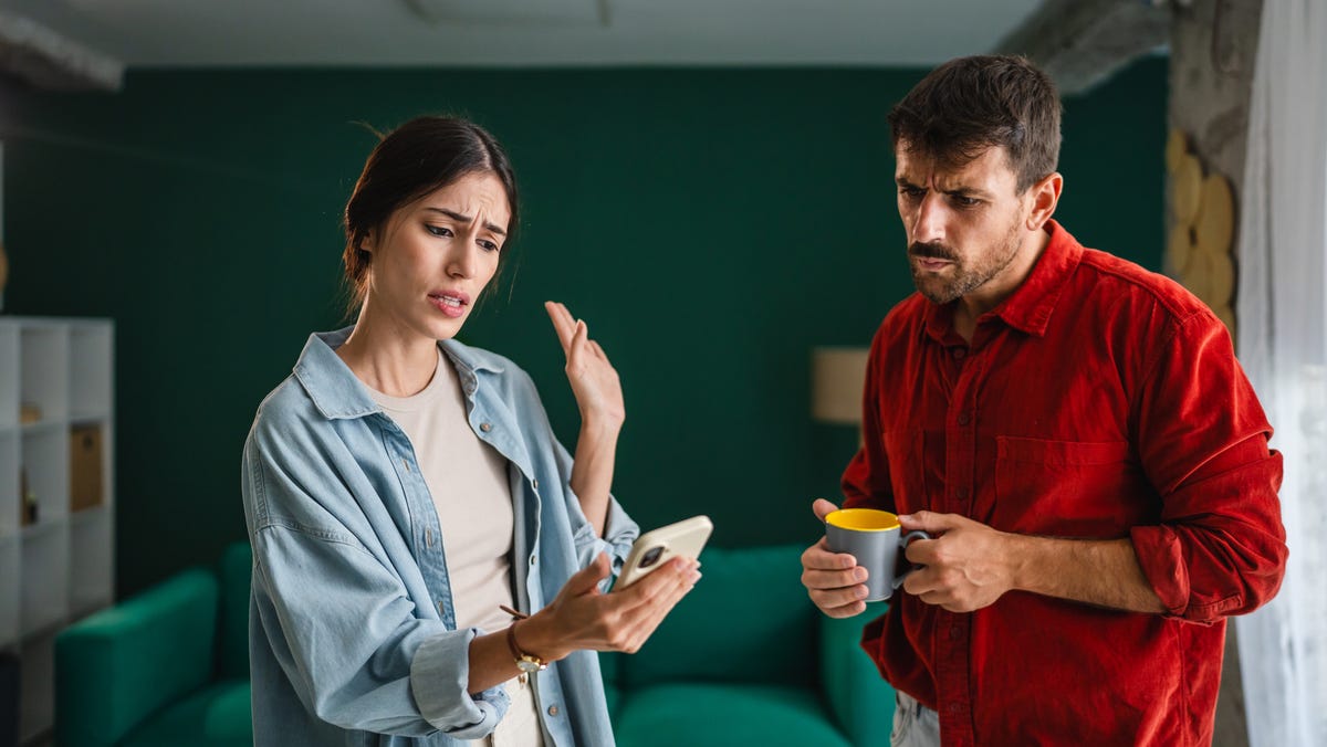 Young couple having a heated discussion in their living room, holding a smart phone and a coffee cup, expressing frustration and disagreement in a tense domestic situation
