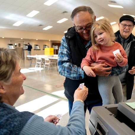 Keith Sjostrand holds his granddaughter, Ila Barrett, 2, while poll worker, Kathleen Forman hands her a voting sticking. Behind them Ila's great-grandfather, Donald Sjostrand, watches with a smile.The family voted at Licking County Church of God in Newark, Ohio, Nov 4, 2025.