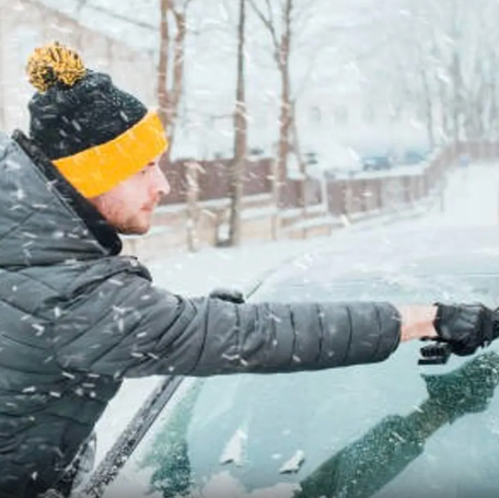 A man uses an ice scraper to remove snow from the windshield of a car.