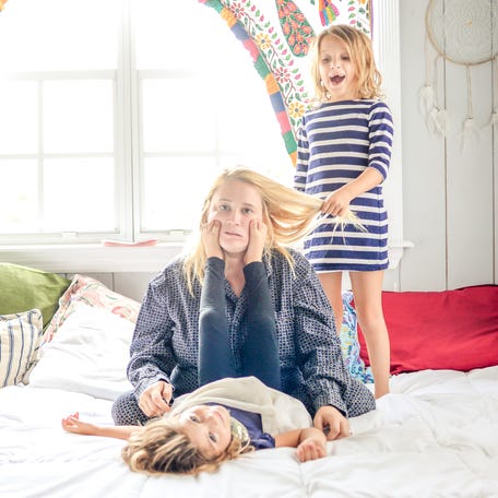 Mother in pajamas with coffee sits on the bed while children jump all around her