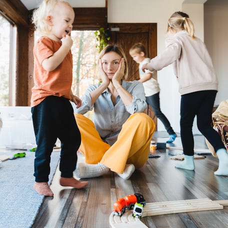 Stressed out mother sitting on floor in middle of toys while children run around.