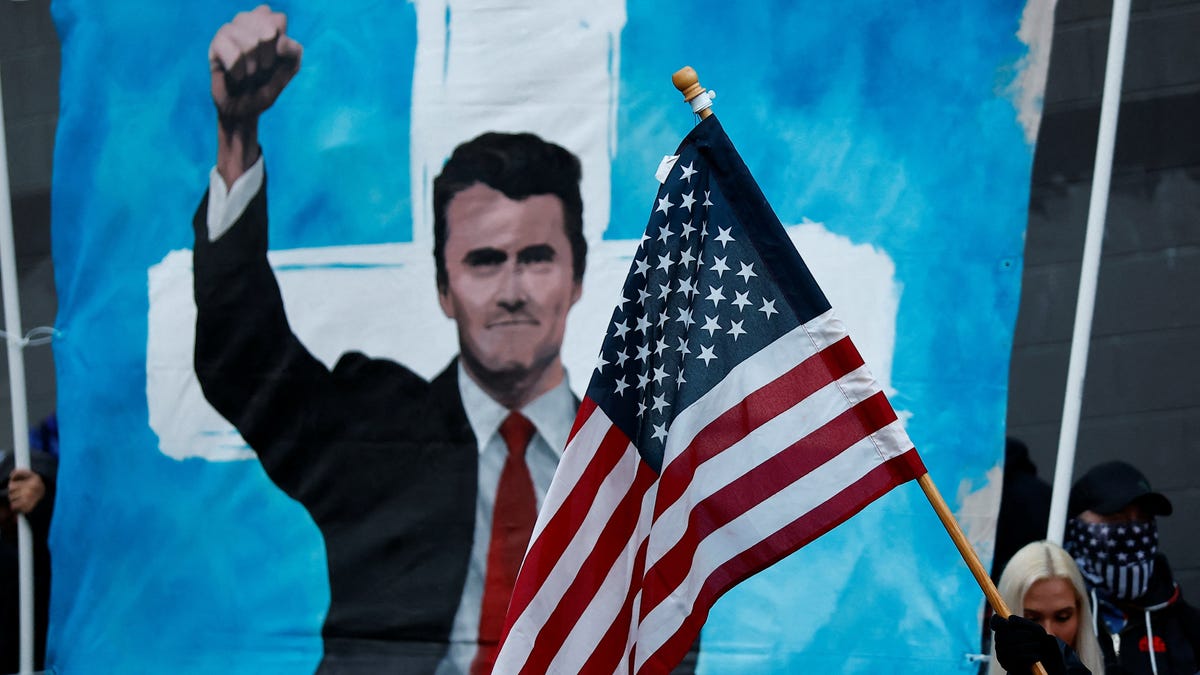 A counter-protester waves a U.S. flag in front of a banner with a picture of slain conservative commentator Charlie Kirk outside an ICE facility during a "World Naked Bike Ride" protest against increased activity by U.S. Immigration and Customs Enforcement (ICE) and potential National Guard deployment in Portland, Oregon, U.S., October 12, 2025.