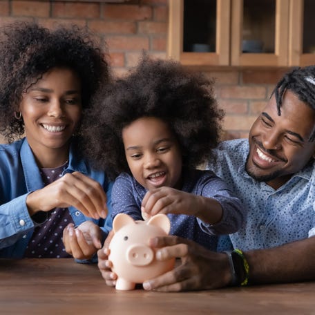Caring young African American couple parents teaching small biracial kid daughter saving money, planning future purchases together, putting coins in small piggybank, financial education for children.