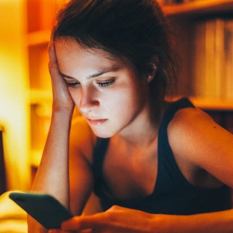 Sad woman sitting alone in a dimly lit room at night, holding her head in one hand while staring at her smartphone screen, looking devastated after being dumped by her boyfriend via text message.