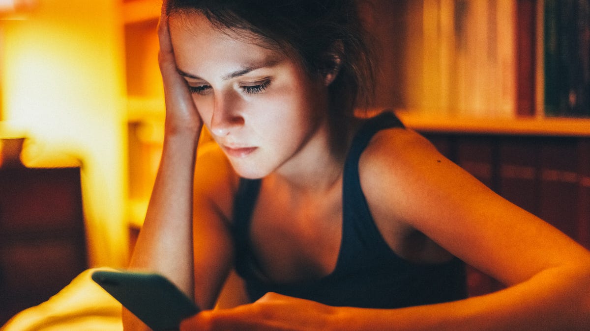 Sad woman sitting alone in a dimly lit room at night, holding her head in one hand while staring at her smartphone screen, looking devastated after being dumped by her boyfriend via text message.