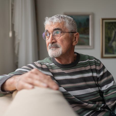 Senior Man Sitting On Sofa By Window And Looking Outside Through Window At Home