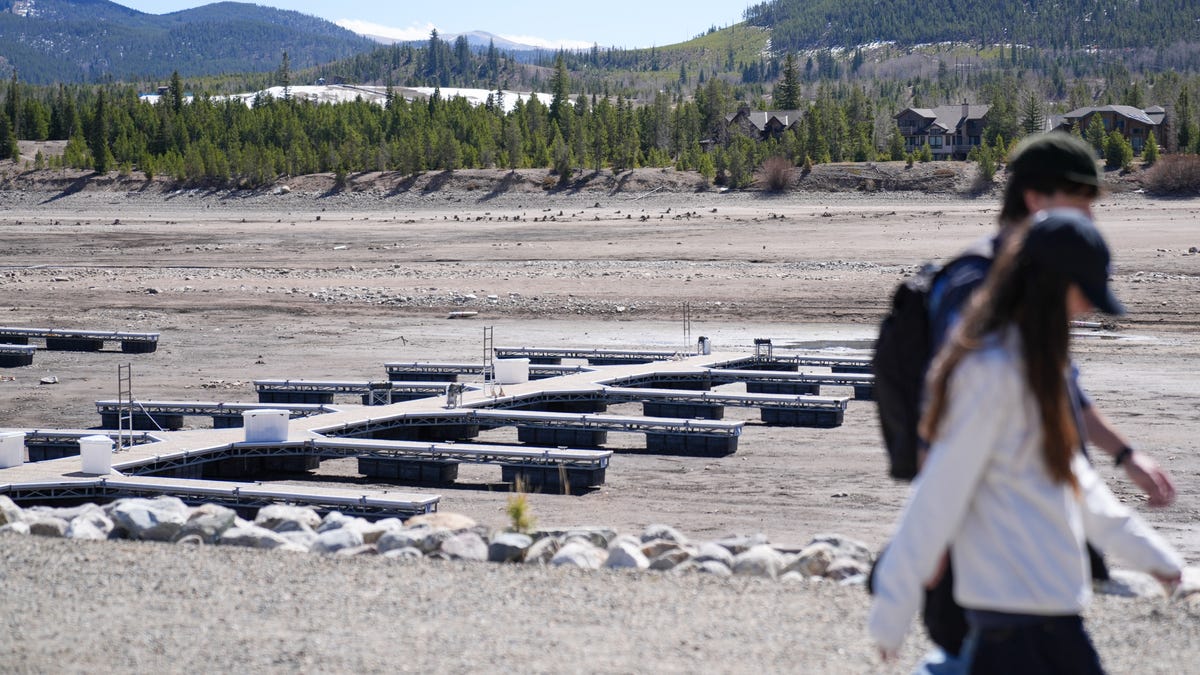 People walk at Lake Dillon in Frisco, Colorado, on March 26, 2026, as lake levels remain low due to poor snowfall during the winter, raising drought concerns. Behind them are docks sitting in mud.