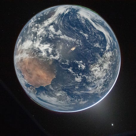 In an image posted by NASA on April 3, 2026, shows a full disk image of Earth, as seen from the Orion capsule. The planet is pale blue, swirling with white clouds and glowing slightly lighter blue in places from reflected light. From the lower left, a large brown landmass is Africa, with the Iberian peninsula twinkling with lights just where the planet curves. In the upper right, aurora glow in a thin green glow, just barely separated from the planet's surface. Earth is set against   the black of space.