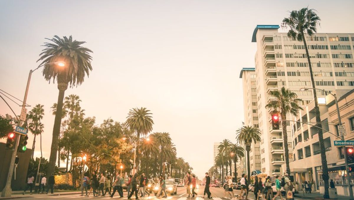 People crossing a city street in Los Angeles that's lined with palm trees.