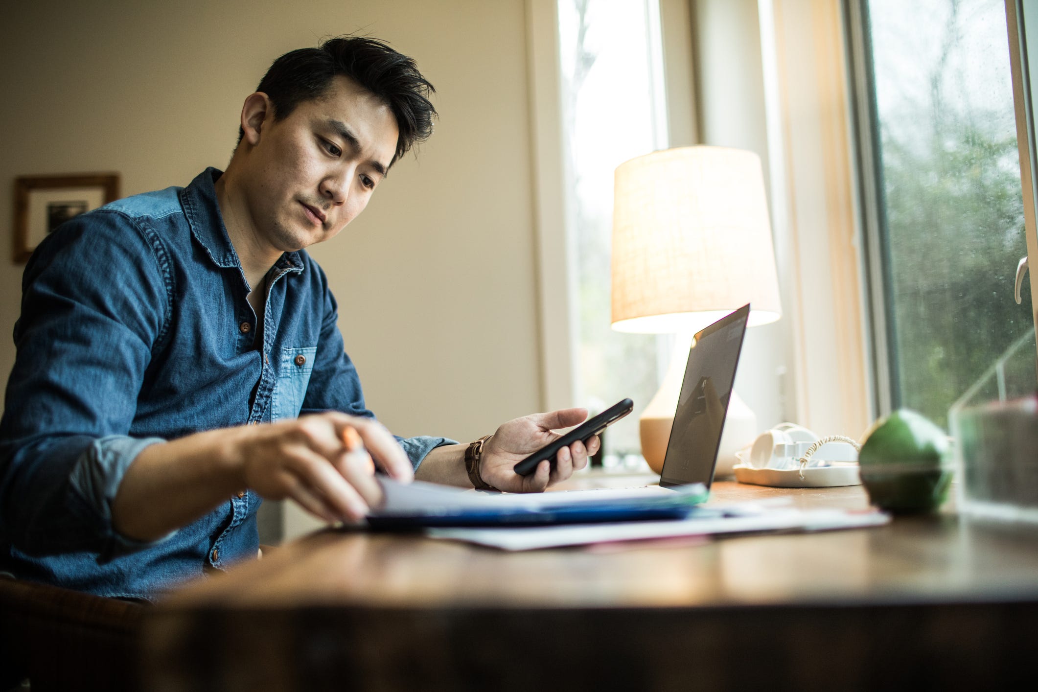 Person holding smartphone and looking at documents on desk.