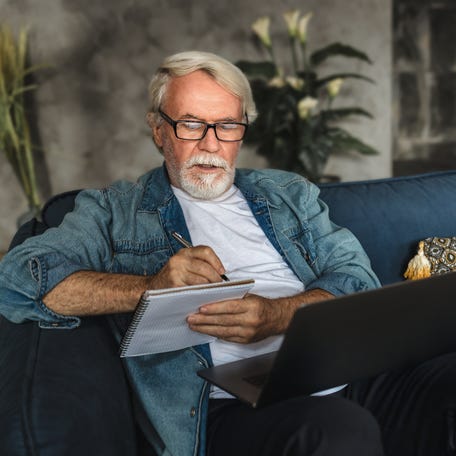 A serious person writing in a notebook while balancing a laptop on their lap.