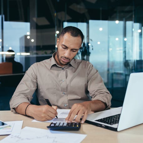 A serious-looking person sits next to a laptop, focusing on a sheaf of papers and a calculator.