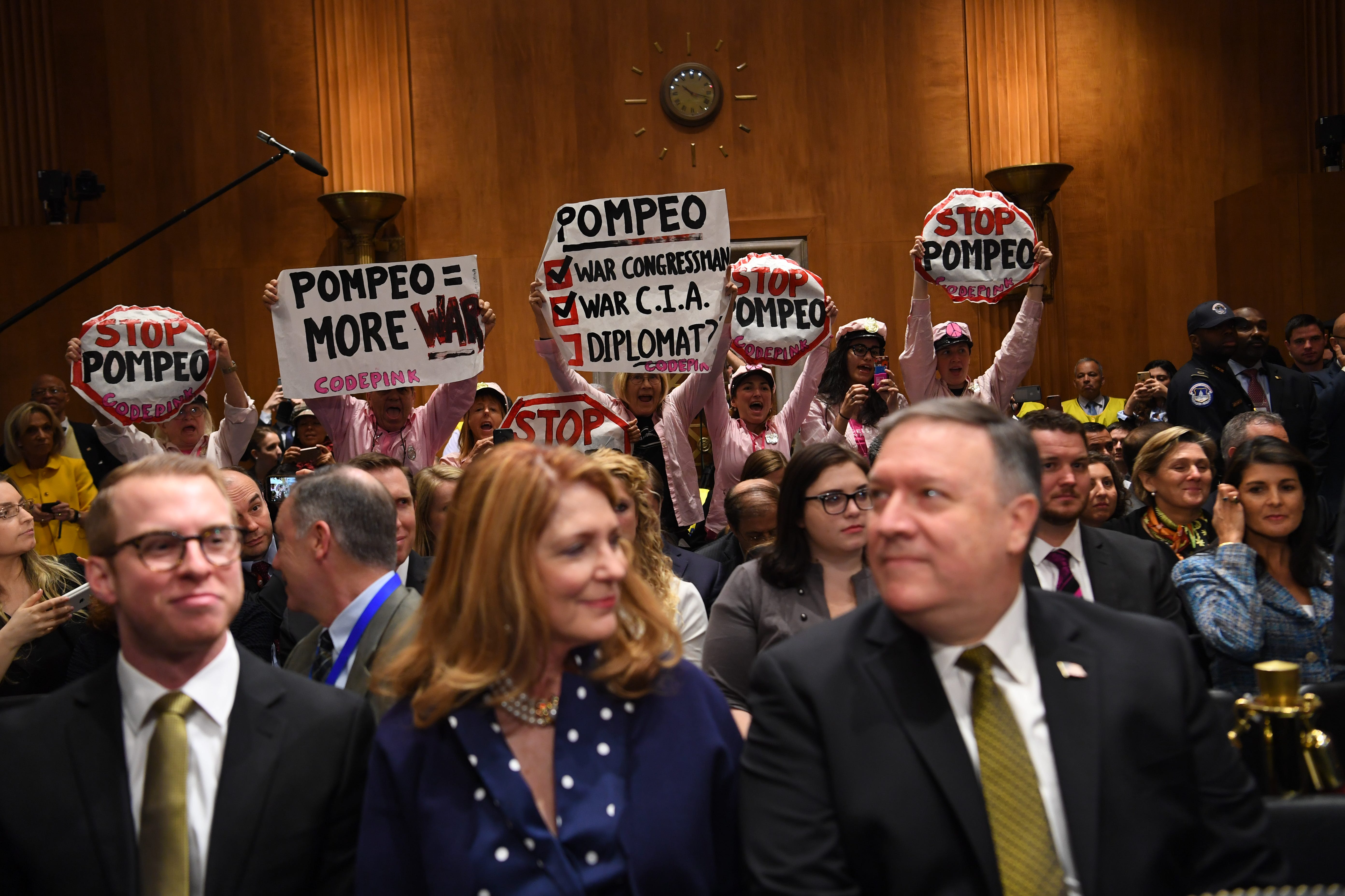 Protestors hold signs from the CODEPINK grassroots anti-war group in the back of the hearing room while Mike Pompeo (R) sits with his wife Susan Pompeo (C) ahead of his scheduled testimony before the Senate Foreign Relations Committee during his conf