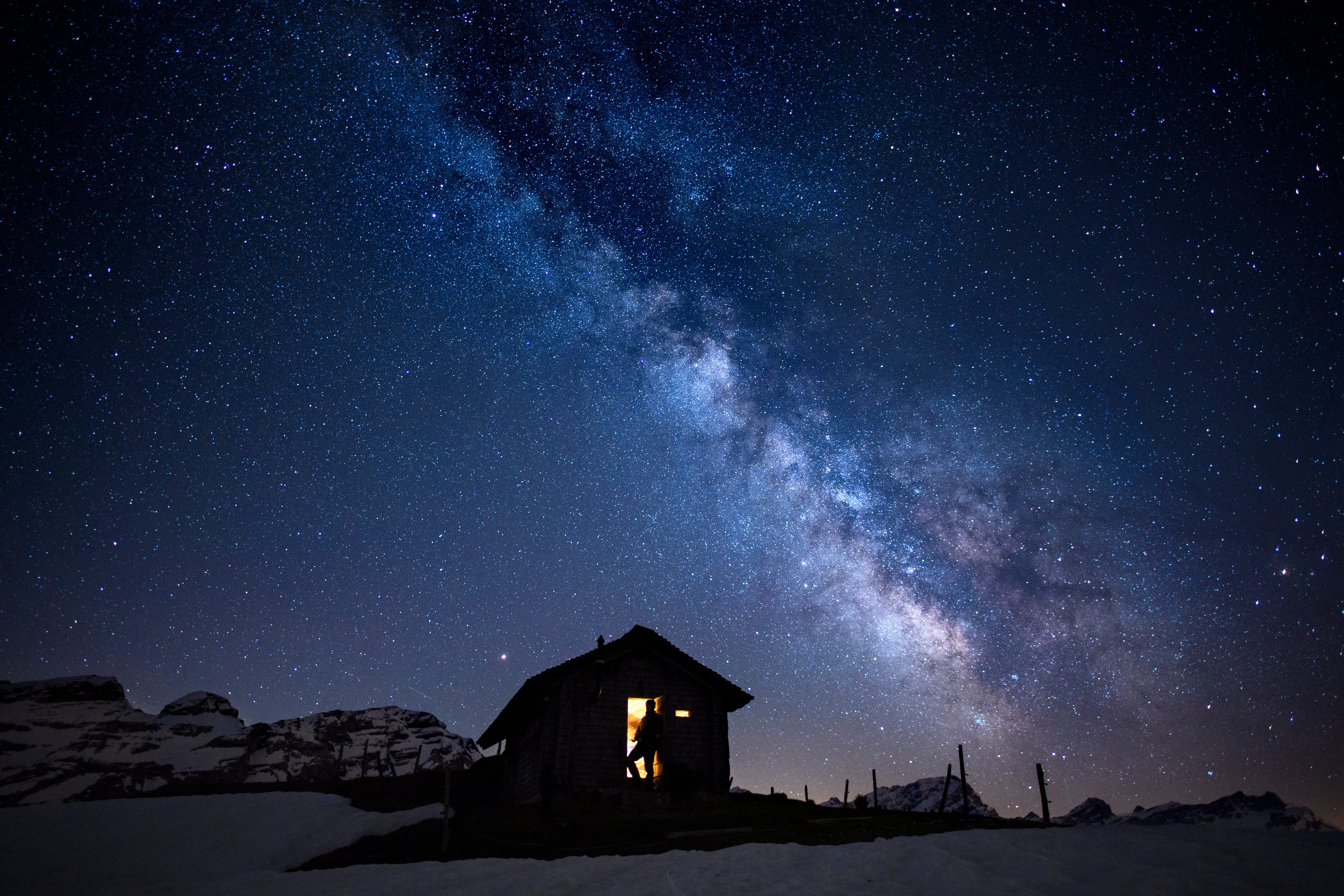 A man is silhouetted against the illuminated inside of a small cabin with a view of the 'Milky Way'  galaxy in the sky over the Ormont Valley, Switzerland.