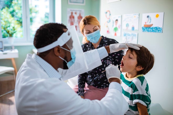 Un profesional médico con equipo de protección tomando un hisopo de mejilla para una prueba de COVID-19 a un niño con otro adulto enmascarado observando.