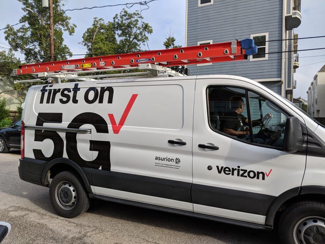 A Verizon maintenance vehicle sits on a roadway near a building. On the side of the van are the words "first on 5G" and a red checkmark.
