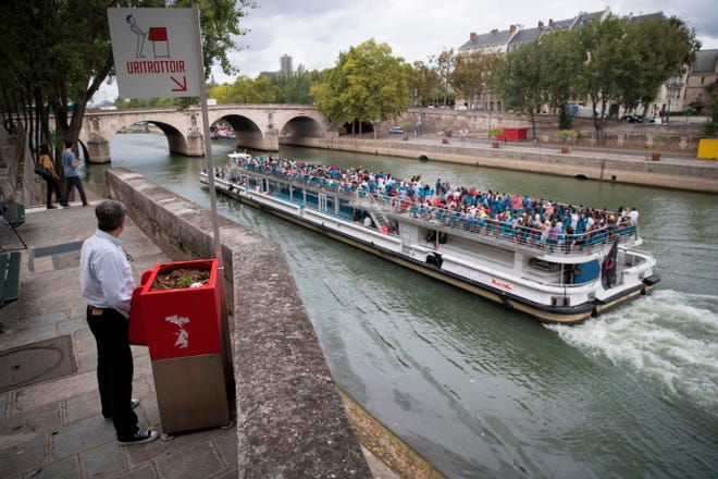 A man stands at a "uritrottoir" public urinal on August 13 on the Saint-Louis island in Paris, as a "bateau mouche" tourist barge cruises past.