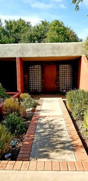 A large oak door framed by equally sized glass block panels and a wall of stacked stone complement one’s arrival.  A colorful palette of potted flowers
bring the beauty of nature into the scene.  A paved sitting area to the left of the home, complete with a sitting wall, beckons conversation and retreat with overhead shelter provided by the roofline provides protection from the elements.