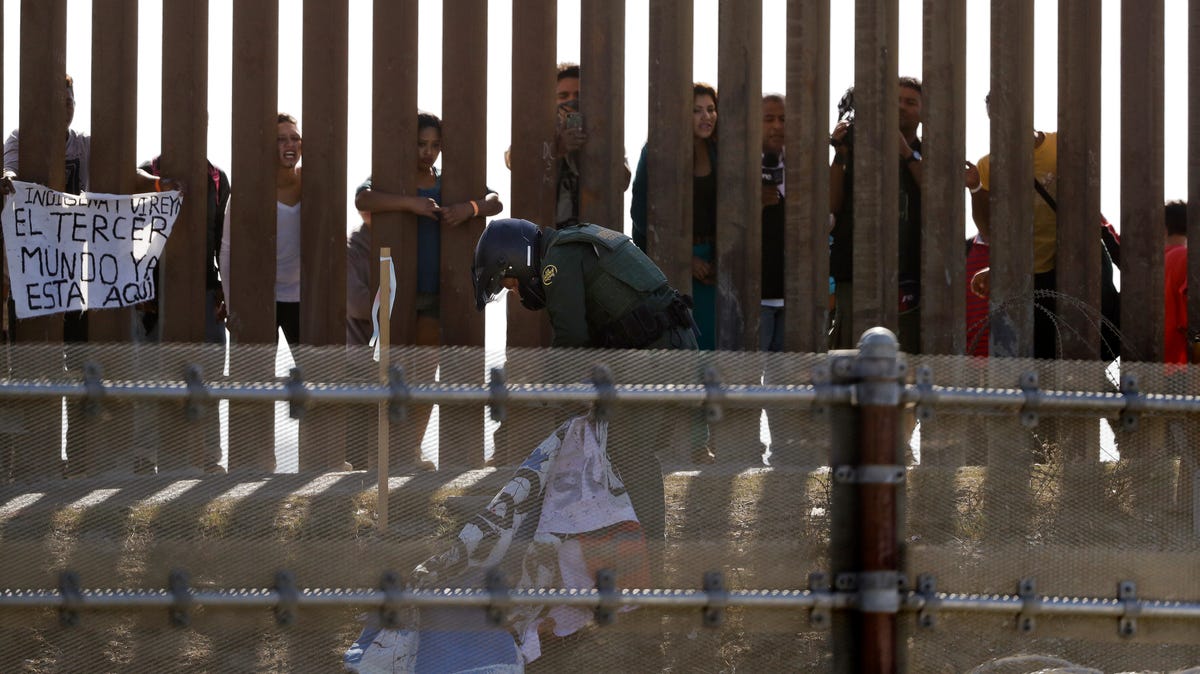 A U.S. Customs and Border Protection officer removes banners posted on a wall at the Mexico-U.S. border, as migrants walk past, as seen from San Diego, Nov. 25, 2018. The banner at left reads: "The Third World is already here." Migrants approaching the U.S. border from Mexico were enveloped with tear gas Sunday after a few tried to breach the fence separating the two countries.